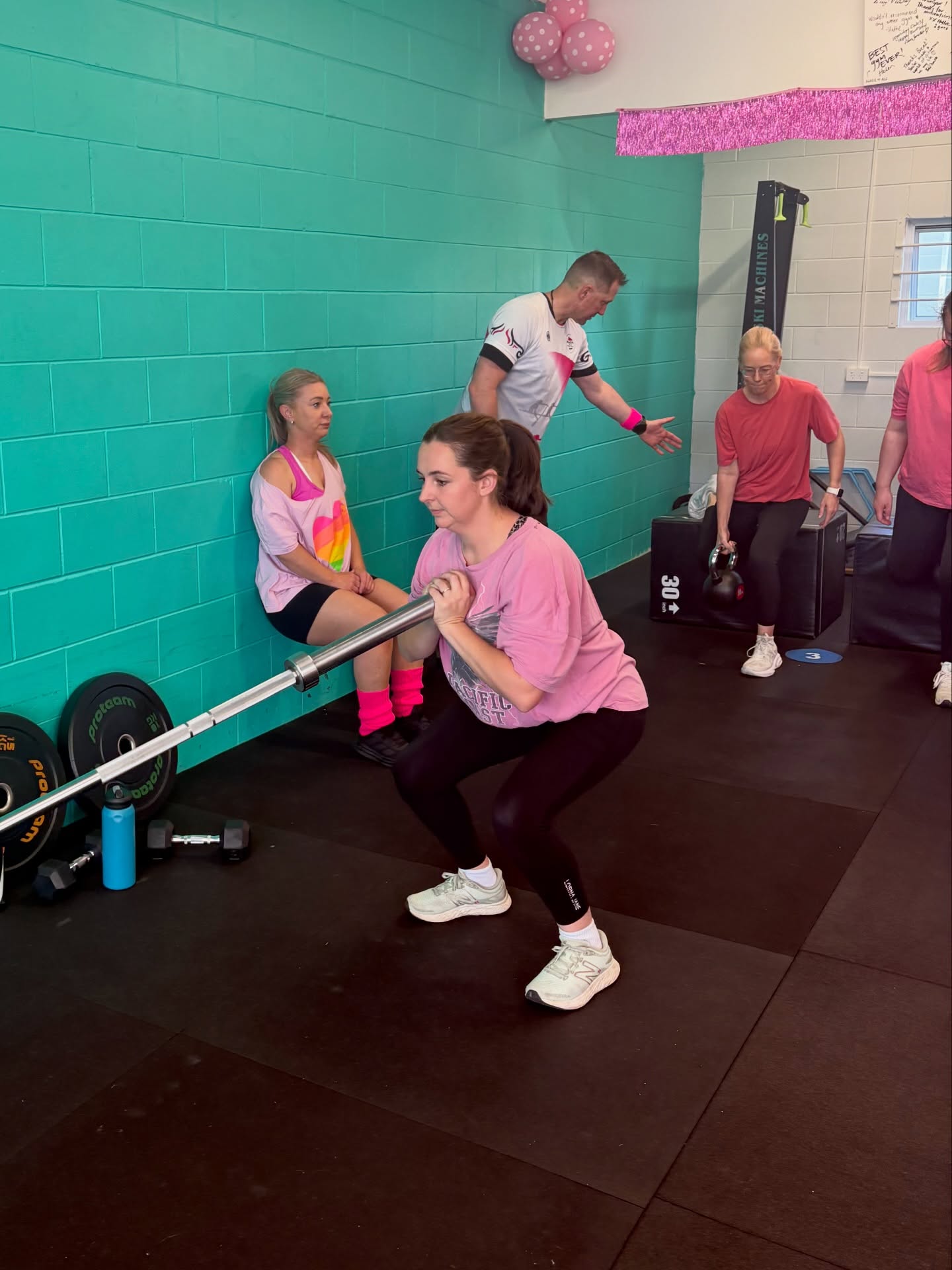 A coach guiding a barbell squat during a fundraiser class, the room decked out in pink